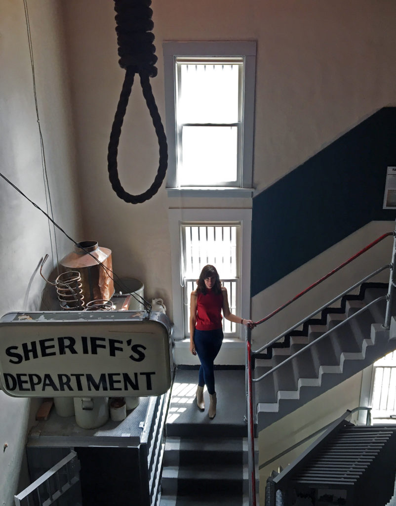 Author Kelly Hartman poses on a stairwell amongst an antique sheriff's department sign and a noose. 