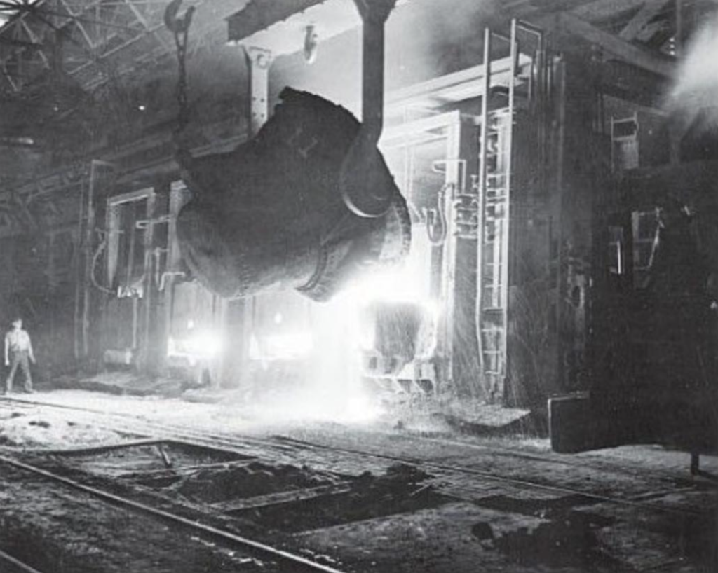 Image of a large vat of iron being poured into a heath furnace as a steel worker stands at a safe distance